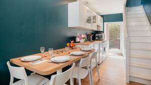Een eetgedeelte met een houten tafel voor vier personen, witte stoelen, een keuken op de achtergrond en een trap naar een buitenruimte.