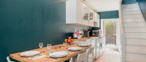 Un comedor con una mesa de madera dispuesta para cuatro personas, sillas blancas, una cocina al fondo y una escalera que lleva a un espacio exterior.