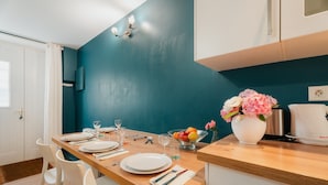 Een eethoek met een houten tafel gedekt voor twee personen, witte stoelen, een vaas met bloemen, een schaal met fruit en een koffiezetapparaat.