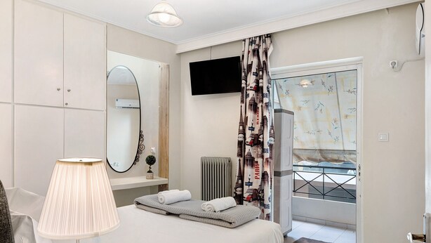 A hotel room with a bed, a bedside lamp, a flat-screen TV, a radiator, a window with patterned curtains, and a door leading to a balcony. - Apartment 'Pallada' with Balcony, Wi-Fi and Air Conditioning (Athina)