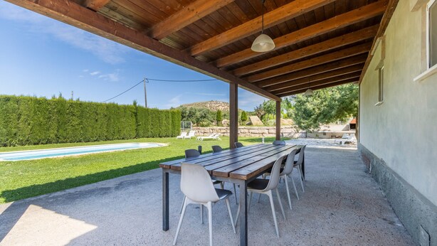 Eine überdachte Terrasse im Freien mit einem Holztisch und -stühlen, einem Schwimmbecken im Hintergrund und einer Steinmauer.
