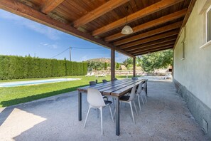 Un porxo exterior cobert amb una taula i cadires de fusta, una piscina al fons i un mur de pedra.