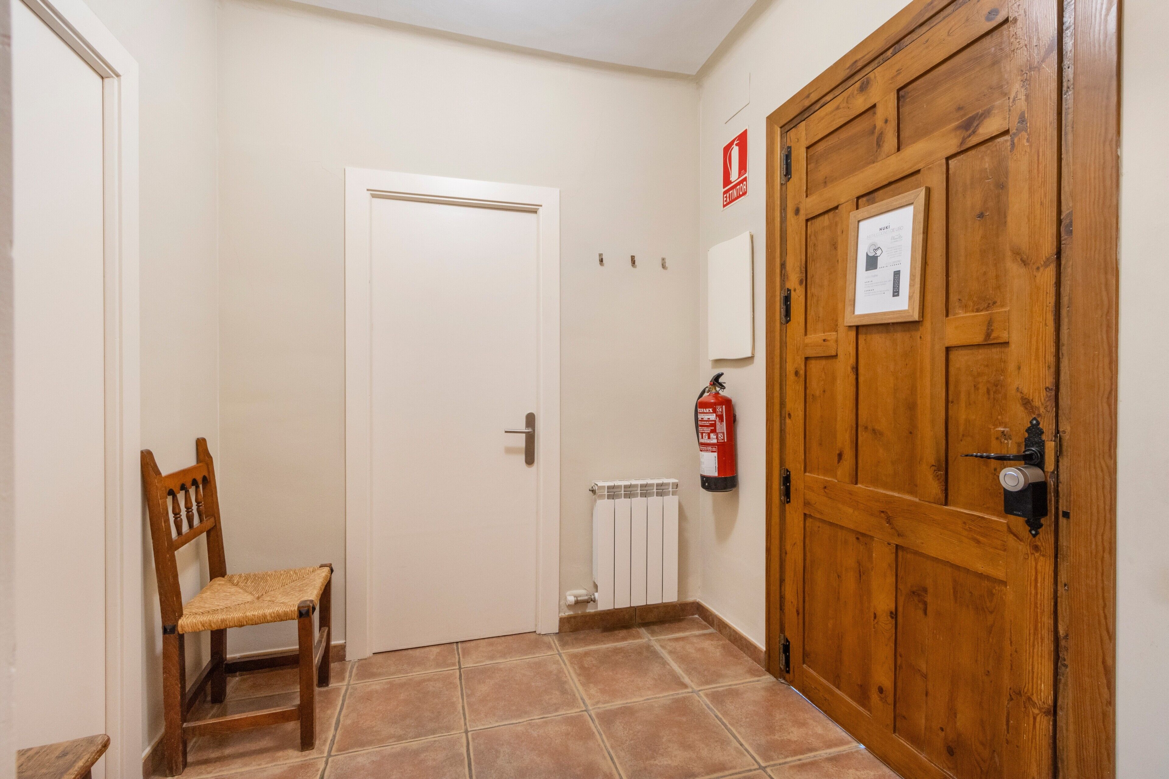Een gang met een houten deur, een witte radiator, een brandblusser en een houten stoel.