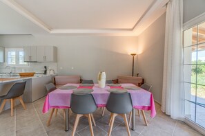 En matplats med ett bord dukat för fyra personer, ett kök i bakgrunden och ett stort fönster med gardiner.