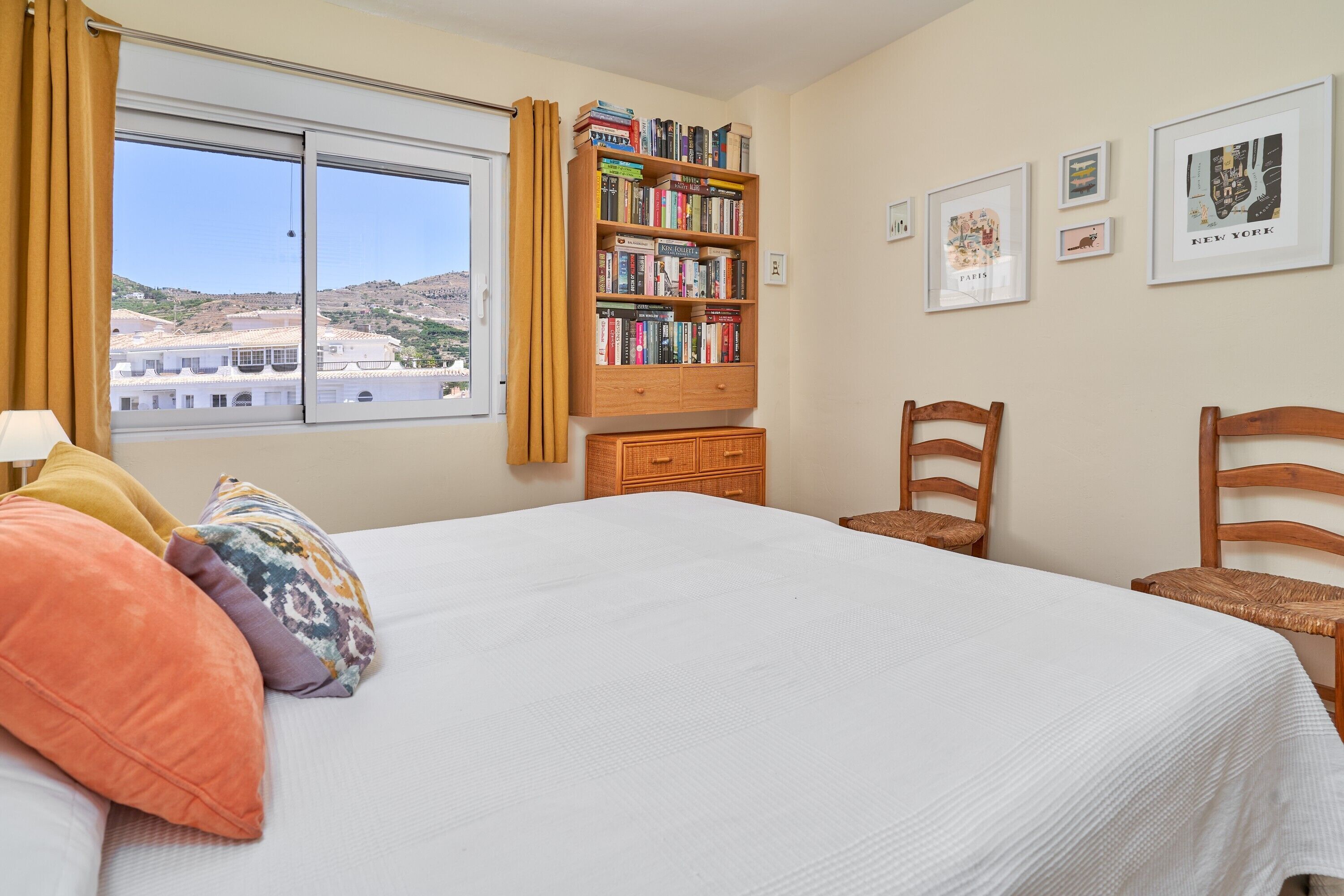 Une chambre avec un grand lit, une bibliothèque, deux chaises, une fenêtre donnant sur des immeubles et un mur orné de tableaux.