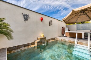 Une piscine à l’eau claire, un mur décoré de motifs représentant des taureaux et des chevaux, un luminaire rouge en forme de cœur et un parasol beige.