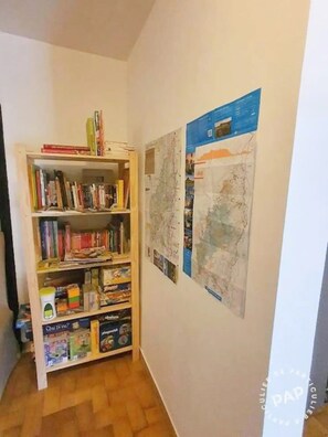 Een gang met een boekenkast vol boeken en gezelschapsspellen, een muur met twee grote kaarten, en een houten vloer.