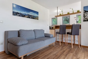 Un salon moderne avec un canapé gris, un plancher en bois et un coin cuisine doté de tabourets de bar et d’une fenêtre.