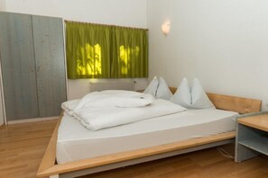 Un lit simple avec un cadre en bois et du linge de lit blanc, une petite table de chevet, un radiateur et une fenêtre avec des rideaux verts.