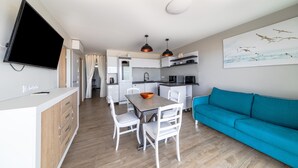 A modern kitchen with white cabinets, a dining table with chairs, a blue sofa, and a flat-screen TV mounted on the wall. - Apartment 'Résidence Le Rêve' with Shared Pool, Wi-Fi and Air Conditioning (La Tranche-sur-Mer)