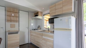 A compact kitchen with wooden cabinets, a white refrigerator, a kettle, and a window with curtains. - Mobile Home 'Mobil-home' with Shared Pool and Private Terrace (Angles)