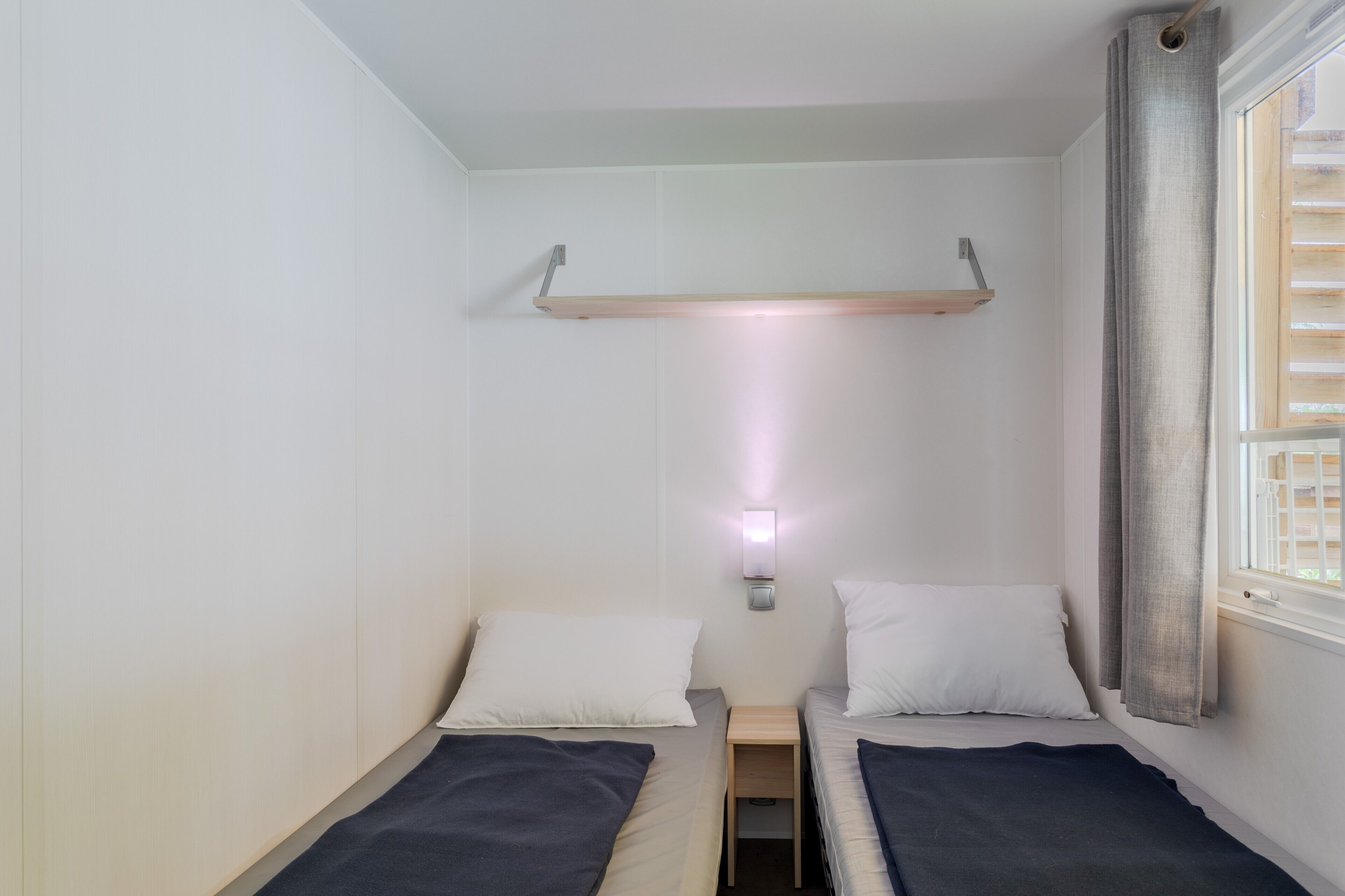 Eine kleine, einfache Unterkunft mit zwei Einzelbetten, einem Nachttisch, einem an der Wand befestigten Regal, einem Fenster mit Vorhängen und einer Wandleuchte.