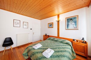 A bedroom with a wooden ceiling, a bed with a green patterned bedspread, a black chair, a wooden nightstand, and framed pictures on the walls.