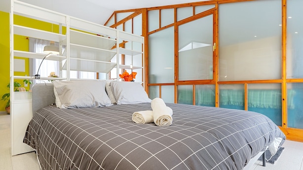A bedroom with a bed, a white wardrobe, a desk, and a large window with wooden frames.