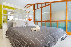 Un dormitorio con cama, armario blanco, escritorio y un amplio ventanal con marcos de madera.