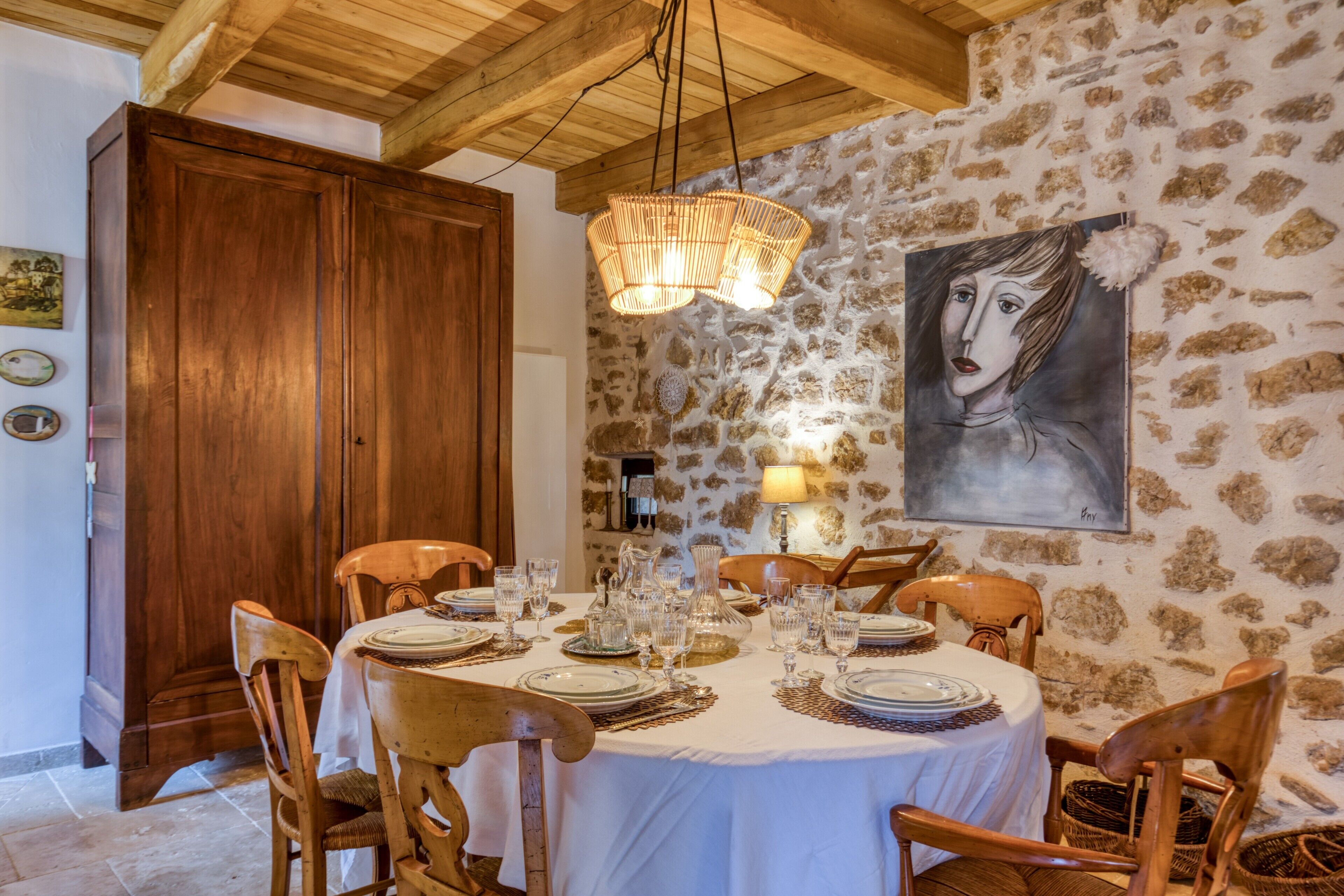 Uma sala de jantar com uma mesa redonda posta para seis pessoas, cadeiras de madeira, uma parede de pedra, teto de madeira e um quadro na parede.