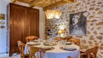 Uma sala de jantar com uma mesa redonda posta para seis pessoas, cadeiras de madeira, uma parede de pedra, teto de madeira e um quadro na parede.