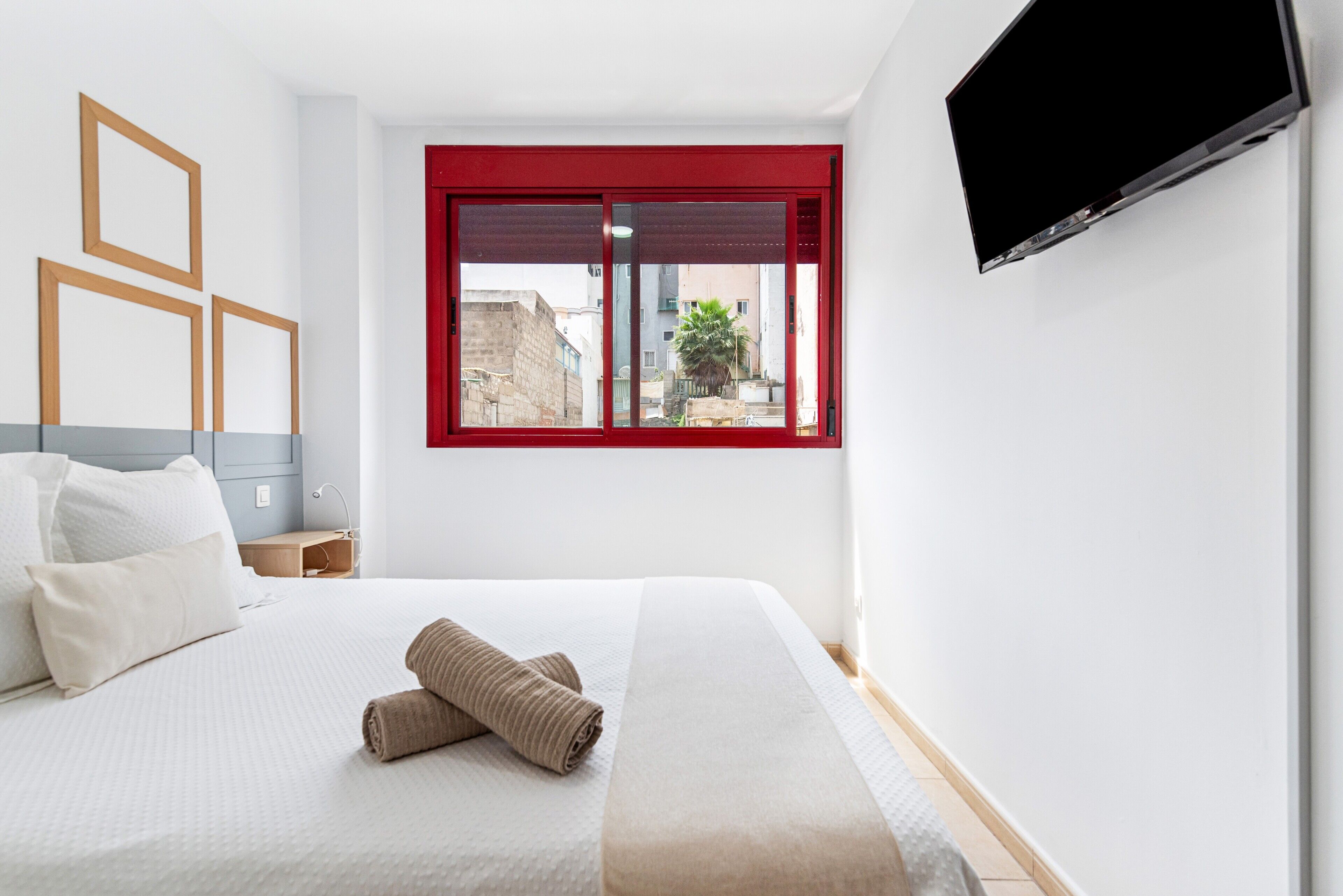 A hotel room with a bed, a TV mounted on the wall, and a window with a red frame.