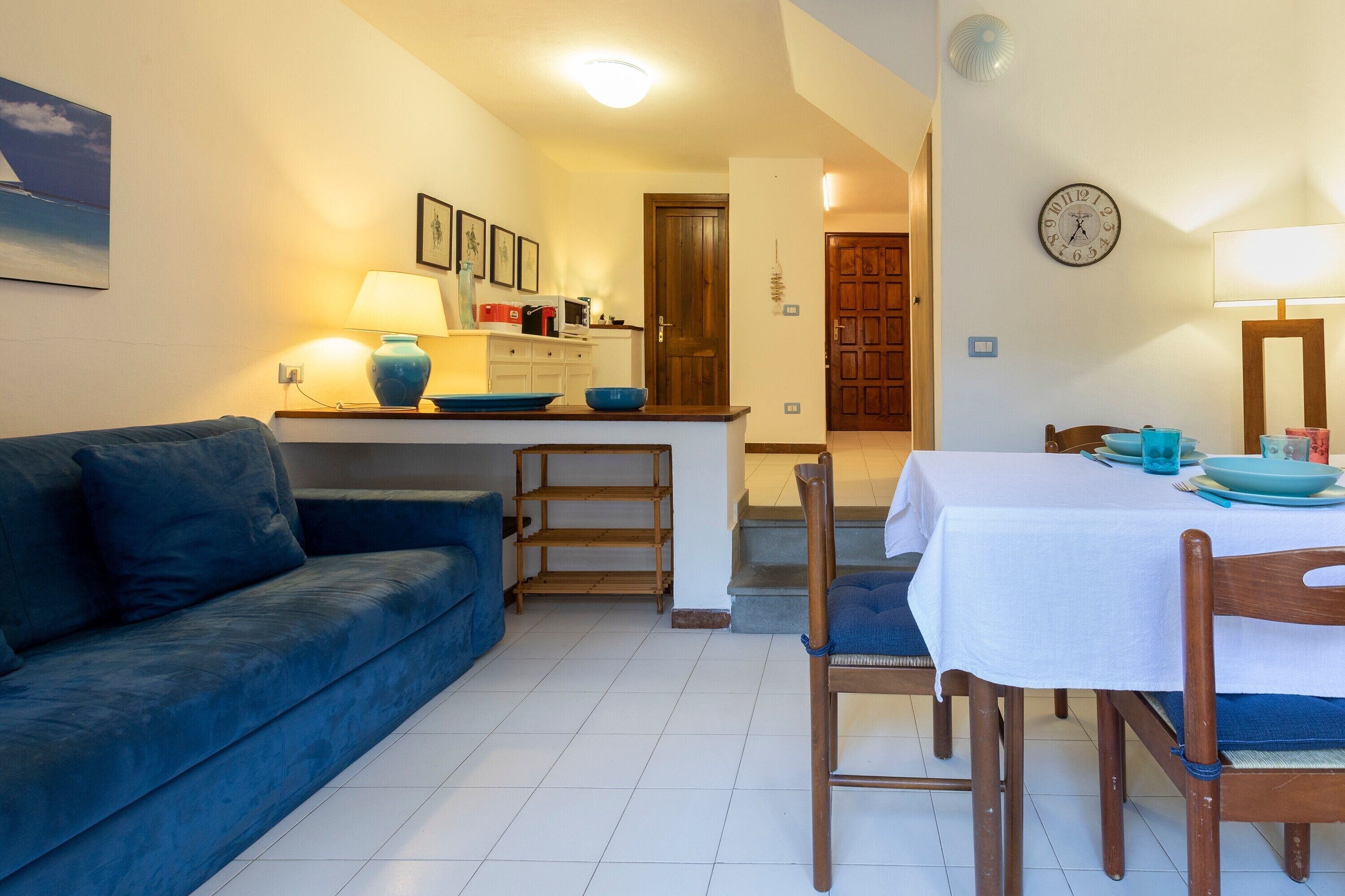 Una sala de estar con sofá, una mesa de comedor con sillas, una zona de cocina con mostrador y armarios, y una escalera que sube a un piso superior.