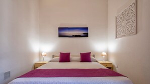 A neatly made bed with a white and purple bedspread, two red pillows, and a nightstand with a lamp. - Holiday Home 'Casetta Al Mare' with Wi-Fi – A/C in Bedroom (Valledoria)