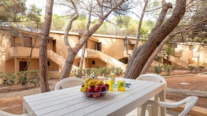 A table with a fruit bowl, a bottle, and glasses, surrounded by white chairs, set outdoors with trees and buildings in the background. - Holiday Home 'Casetta Al Mare' with Wi-Fi – A/C in Bedroom (Valledoria)