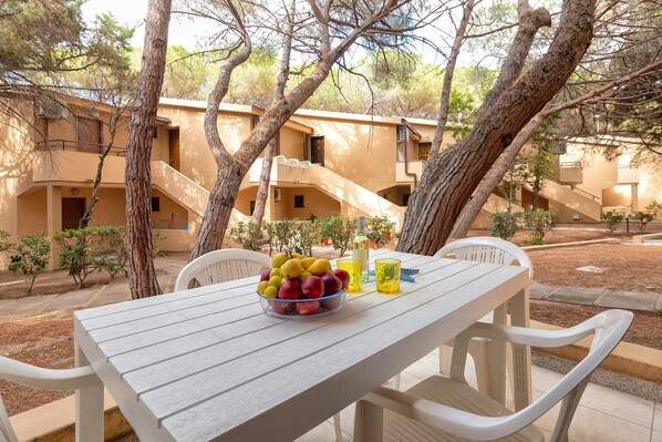 A table with a fruit bowl, a bottle, and glasses, surrounded by white chairs, set outdoors with trees and buildings in the background.