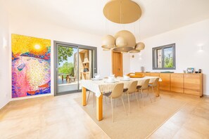 A modern dining area with a table set for four, a large abstract painting on the wall, and a view through the glass door. - Villa 'Viduleto' with Mountain View, Private Pool and Wi-Fi (Felanitx)