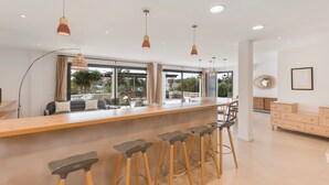 Une cuisine moderne avec un bar en bois, des tabourets de bar, des luminaires suspendus et de grandes fenêtres offrant une vue sur l’extérieur.