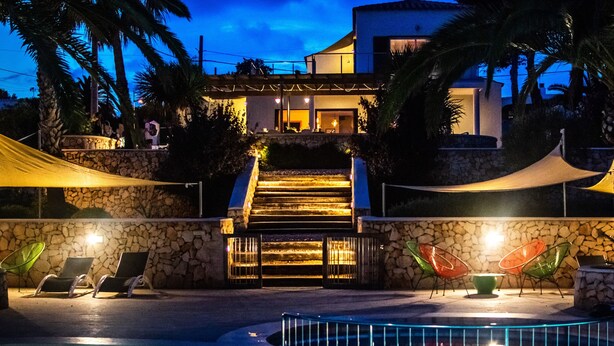 Un espace piscine agrémenté d’un mur en pierre, d’un escalier menant à un bâtiment et de coins salon extérieurs avec des chaises.