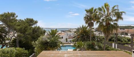 A wooden deck overlooking a swimming pool, surrounded by greenery and buildings in the background.
