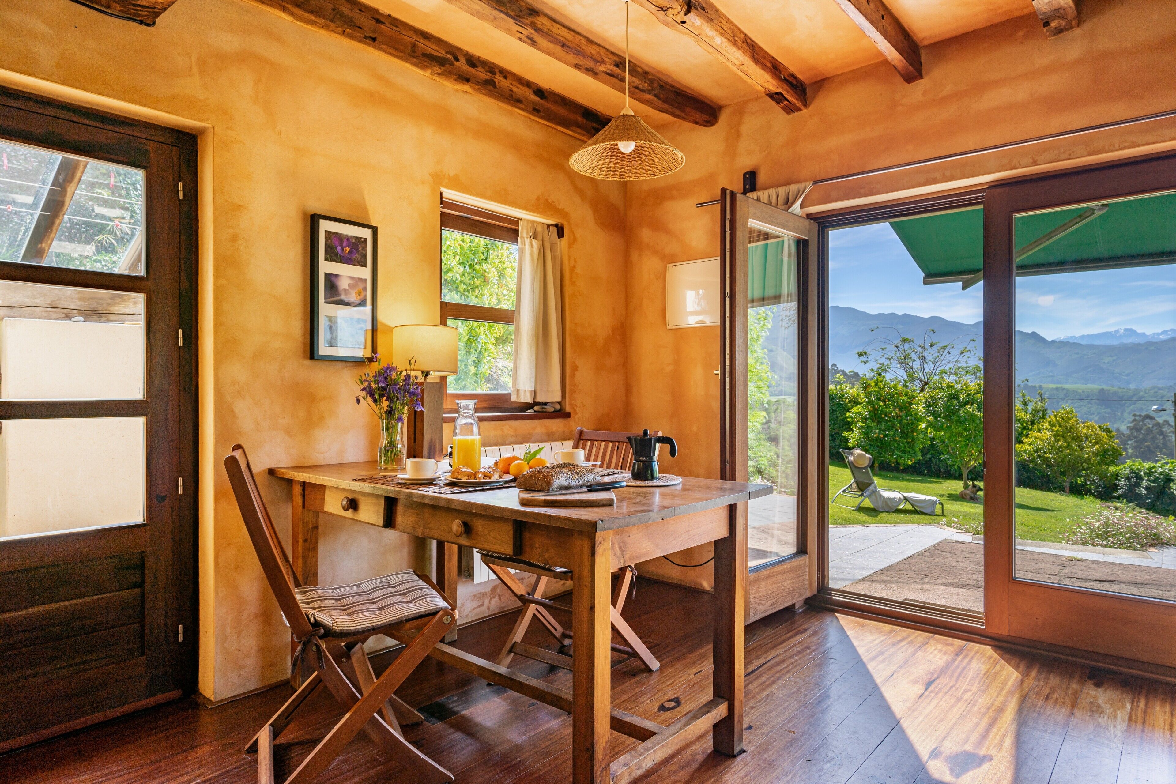 Una mesa de comedor de madera con sillas, una ventana con cortinas y vistas a un jardín y montañas a través de un ventanal.