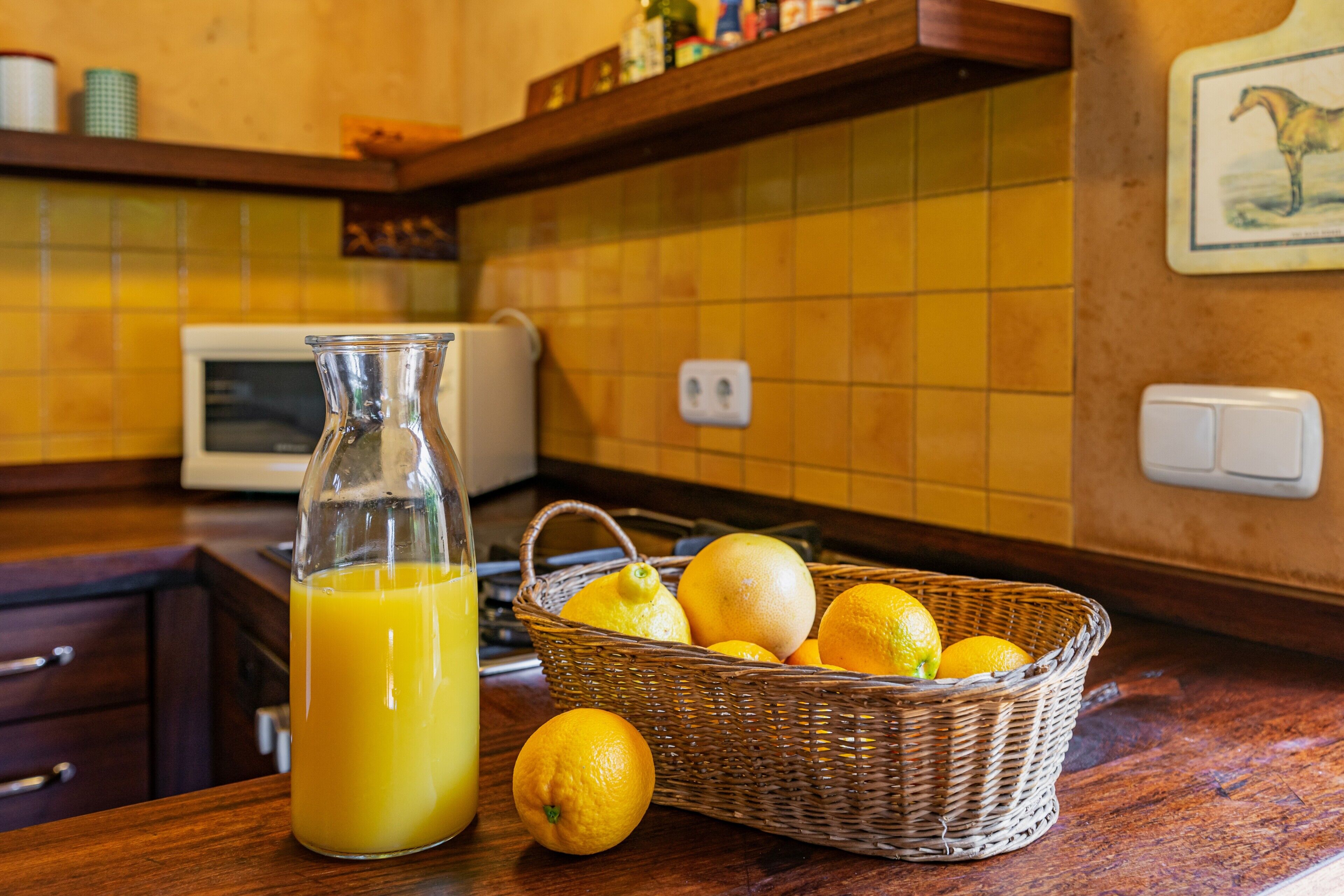 A kitchen with a glass bottle of orange juice, a wicker basket with lemons, and a microwave.