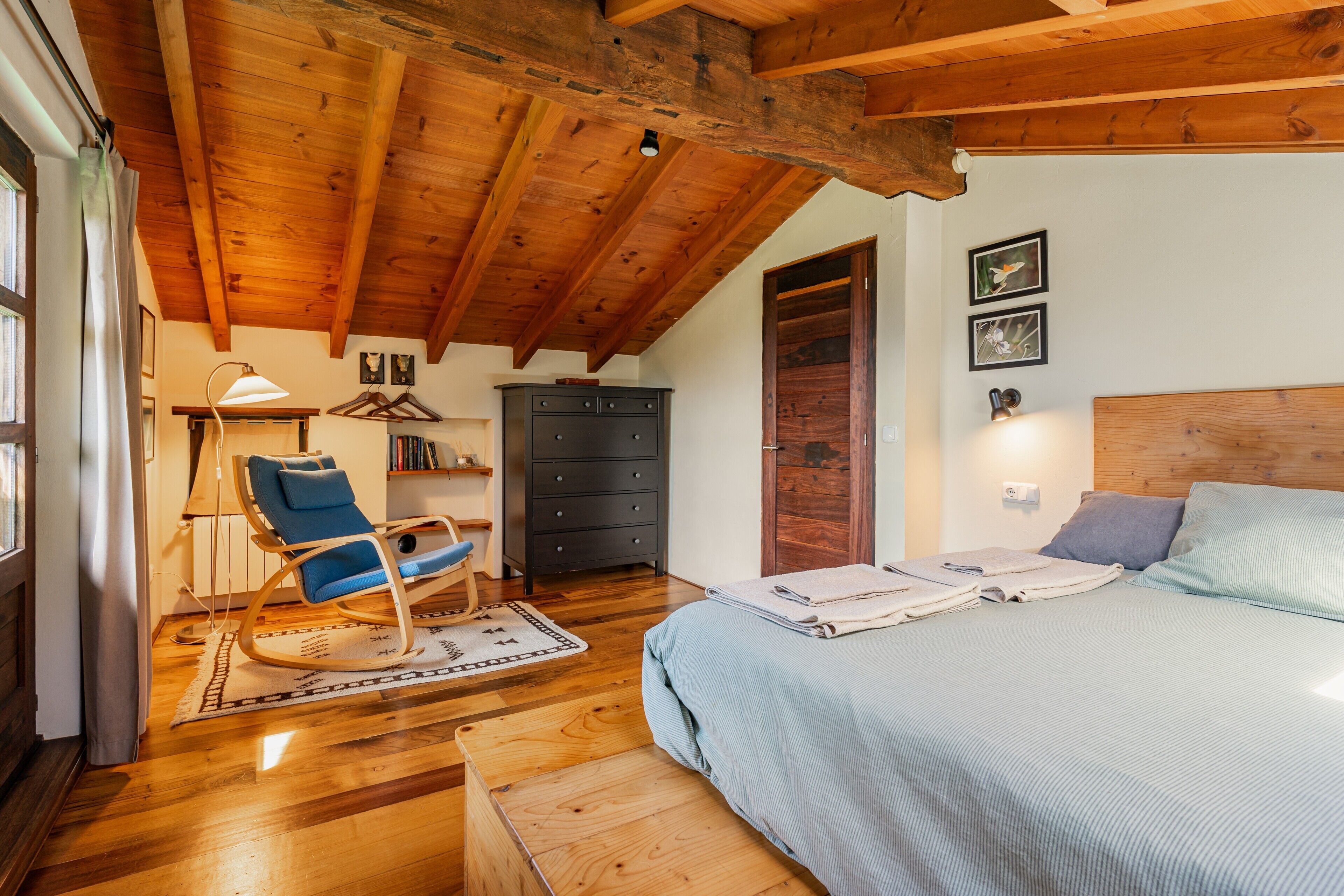 A bedroom with a wooden ceiling, a bed with pillows, a rocking chair, a dresser, and a lamp.