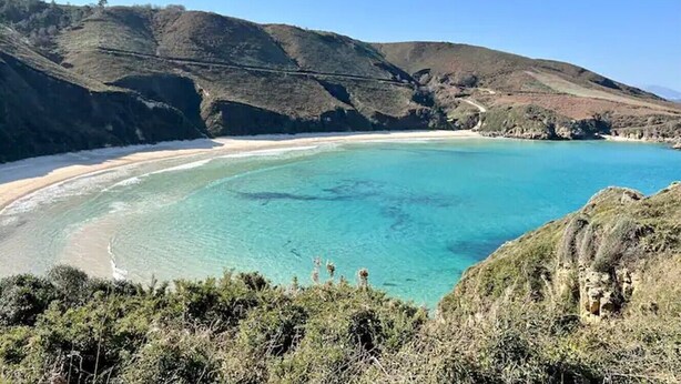 Eine Küstenlandschaft mit Strand, klarem türkisblauem Wasser und hügeligem Gelände.