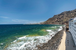A coastal view with a rocky shoreline, clear blue sea, and a building with balconies overlooking the ocean.