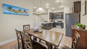A modern kitchen with dark cabinets, a stainless steel refrigerator, and a dining area with a wooden table and chairs. - Entire Home in Denton, Texas. Centrally Located! Luxurious Home Away from Home! (Denton)