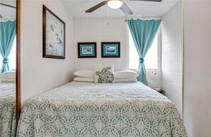 Une chambre à coucher avec un lit, deux tableaux encadrés au mur, un ventilateur de plafond et des rideaux de couleur claire.