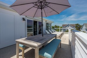 Une table en bois, un banc, un parasol et une vue sur une maison dotée d’un balcon.