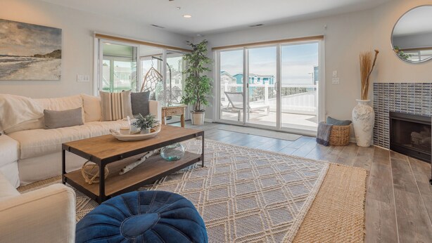 Un salon spacieux avec un canapé, une table basse et un pouf bleu. Une grande fenêtre offre une vue sur une maison et un balcon.