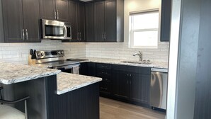 A modern kitchen with dark cabinets, stainless steel appliances, a marble countertop, and a window providing natural light. - Relax on the deck and enjoy the beautiful lake views in Vernon, (North Okanagan) (Vernon)