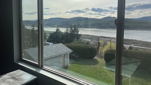 A room with a large window overlooking a lake, hills, and a small house. - Relax on the deck and enjoy the beautiful lake views in Vernon, (North Okanagan) (Vernon)