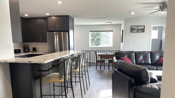 A modern kitchen with dark cabinets, a stainless steel refrigerator, and a marble countertop. There is a dining area with a table and chairs, and a black leather sofa with red cushions. - Relax on the deck and enjoy the beautiful lake views in Vernon, (North Okanagan) (Vernon)