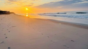 Una playa con huellas que conducen al océano, un muelle de madera y un atardecer al fondo.