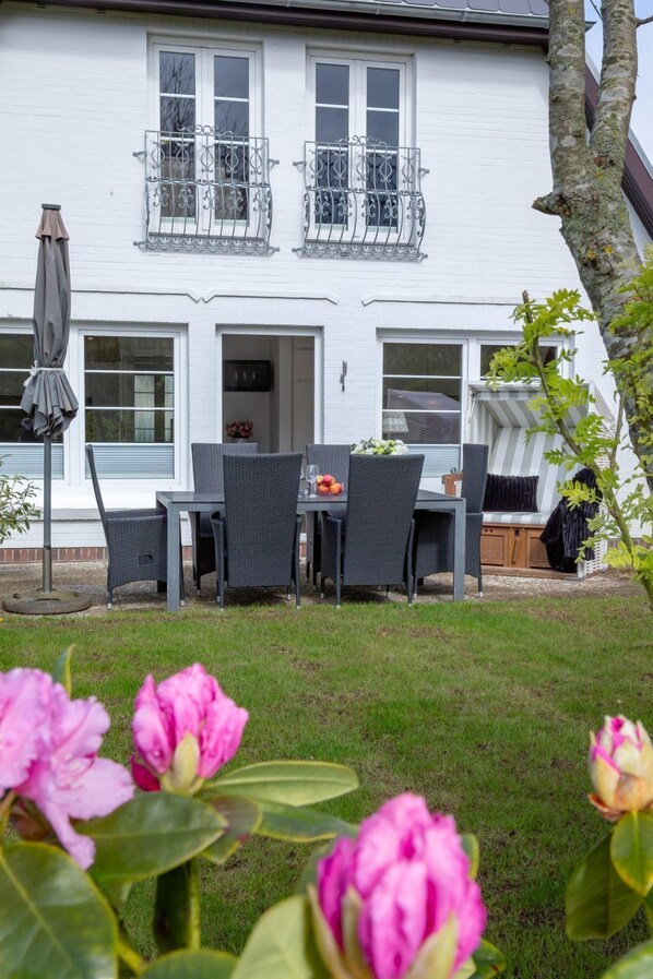 Una casa blanca con balcón, un comedor con mobiliario de mimbre y un jardín con flores rosadas.