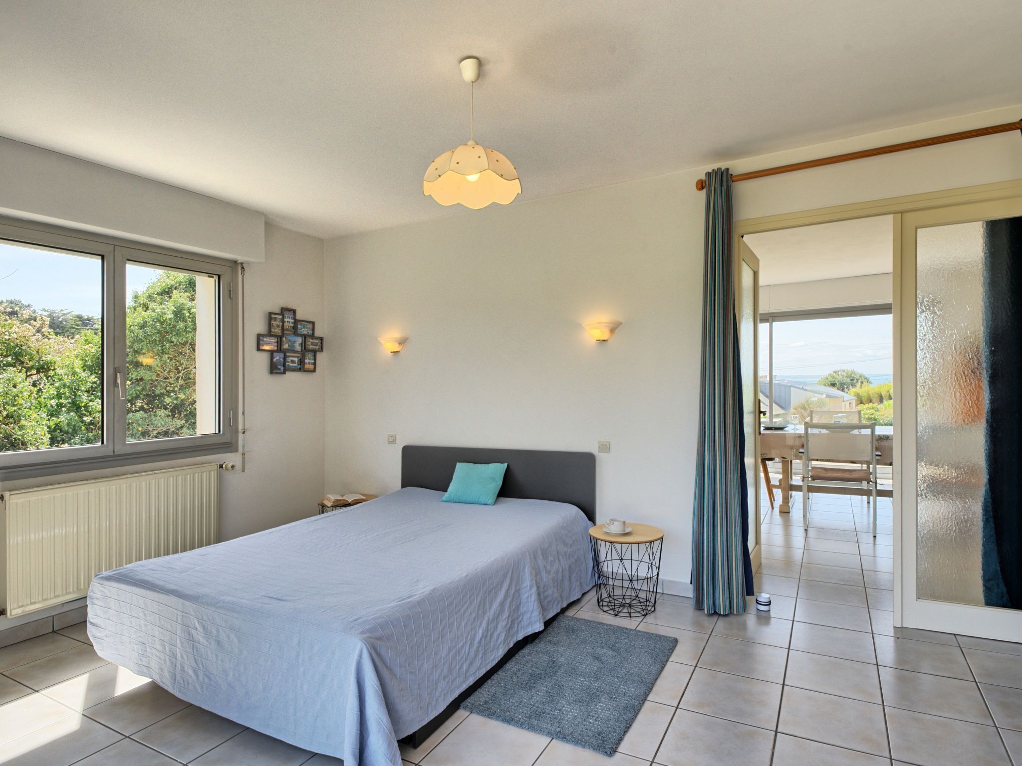 A bedroom with a bed, a small round table, a wall-mounted light, and a view of the outdoors through a window.