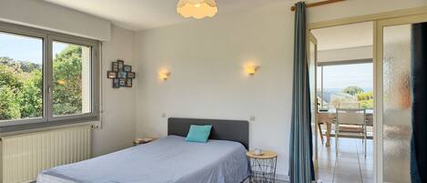 A bedroom with a bed, a small round table, a wall-mounted light, and a view of the outdoors through a window.