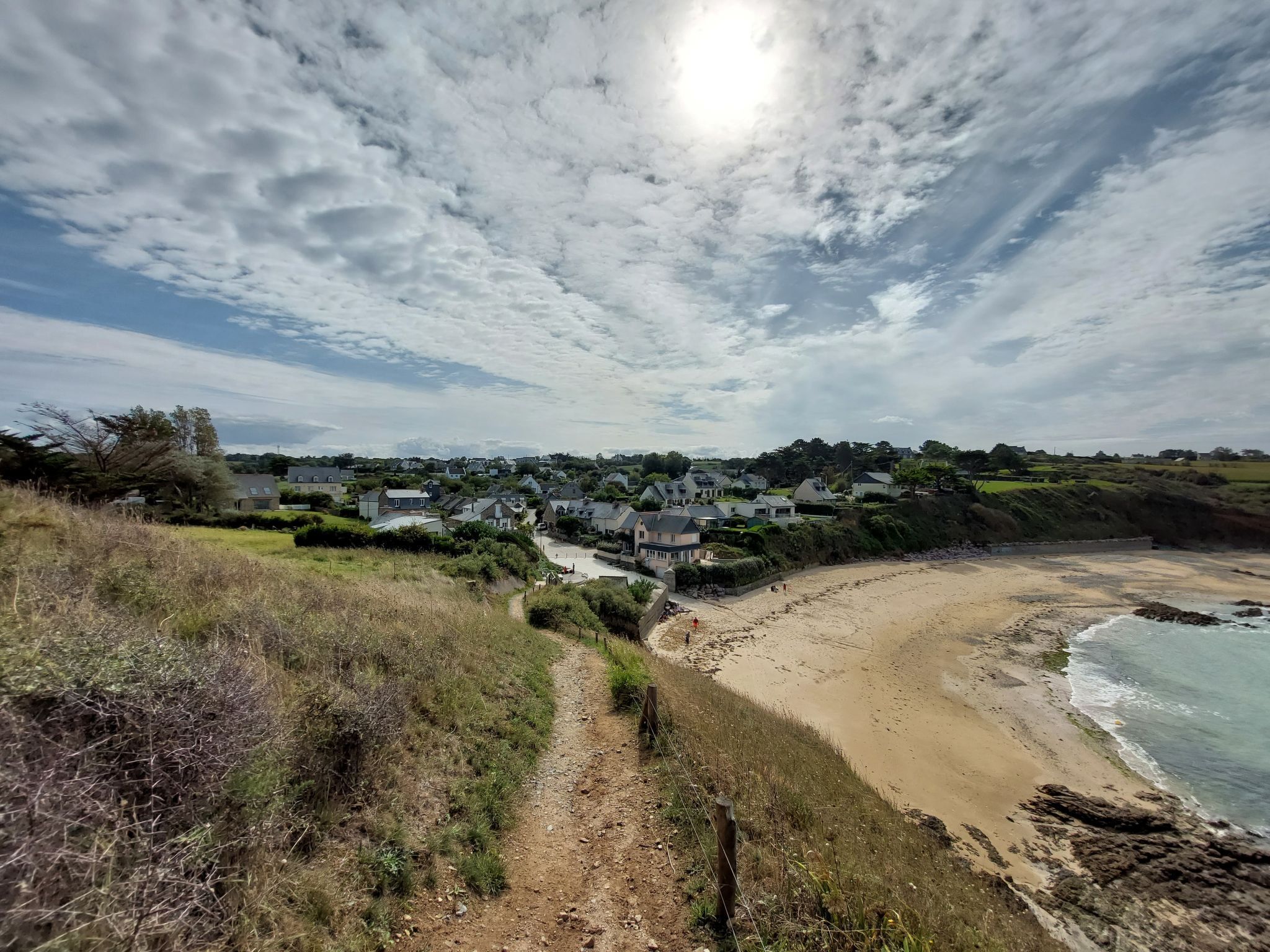 A coastal view with a sandy beach, a path leading to a residential area, and houses with sloped roofs.