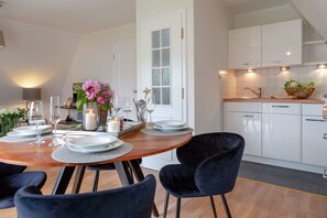 Une salle à manger moderne avec une table en bois dressée pour quatre personnes, des chaises en velours noir et une cuisine dotée de meubles blancs et d’un dosseret carrelé.