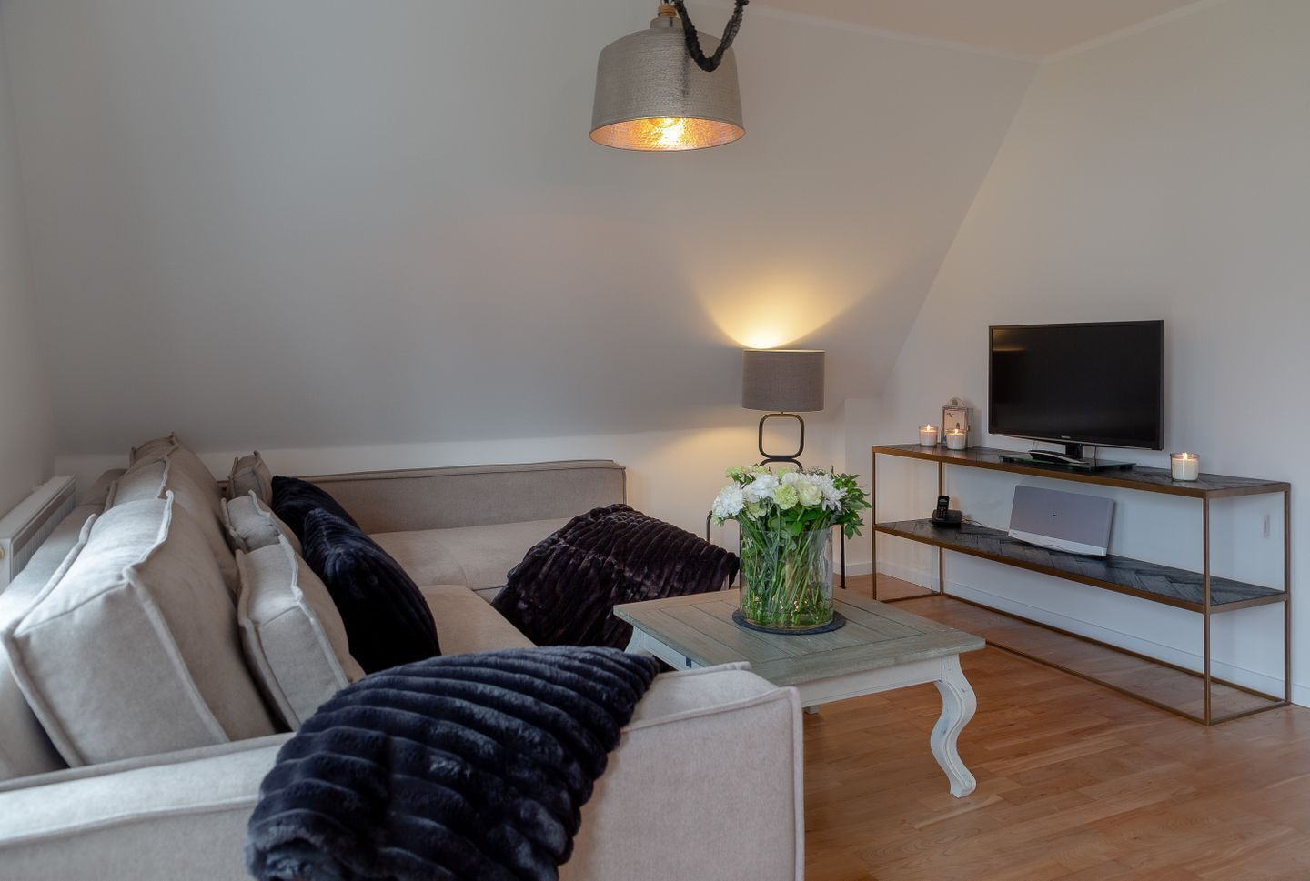 Uma sala de estar moderna com um sofá, uma mesa de centro com um vaso de flores, uma TV sobre um suporte de madeira e uma luminária.