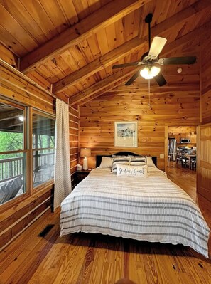 A wooden cabin bedroom with a bed, a ceiling fan, a window, and a dining area visible through an open door.